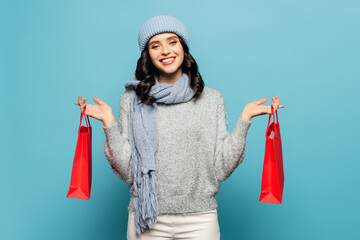 Front view of woman in winter outfit looking at camera while holding red shopping bags isolated on blue