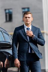 young confident businessman touching tie near car outdoors.