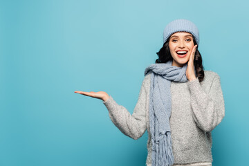 Front view of woman with open mouth, wearing winter outfit and pointing with hand isolated on blue
