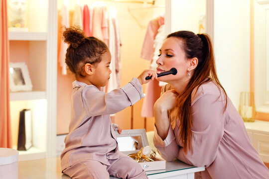 Caucasian mother and African American daughter in pink pajamas in dressing room and bedroom. Brush for cosmetics paint each other