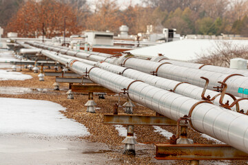 Side view of a pipeline running over snowy terrain near an energy plant. Heavy duty metal pipes carrying gas and oil are supported by columns and base plates. Rusty metal rings encircle pipes.