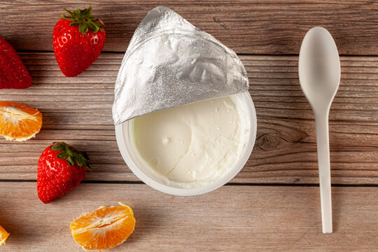 Flat Lay Image Of A Store Bought Plastic Cup Of Plain Low Fat Greek Yogurt With The Aluminum Foil Seal Peeled Off. It Is On Wooden Background With A Plastic Spoon And Fresh Fruits. A Diet Idea Concept