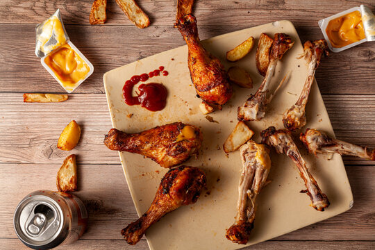 Flat Lay Of An Untidy Wooden Table With A Plate Of Deep Fried Or Grilled Seasoned Chicken Drumsticks With Half Eaten, Leftover Bones, Dipping Sauces In Packs, And Potato Chunks In Background.