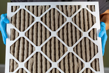 A person wearing blue gloves is holding a heavily clogged dirty air filter in hands before replacing it with the new one. Paper based, card board frame plated filters are used in hvac, ac , furnaces.