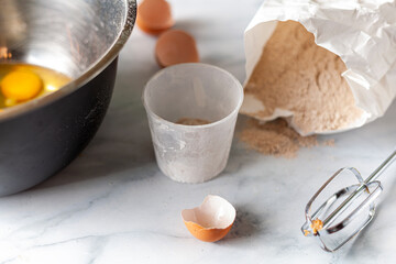 Pastry being prepared on marble kitchen countertop with a paper bag of wholewheat flour, broken egg shells, measuring cup and mixer attachment seen. Raw egg whites and yolks are in a shiny metal bowl