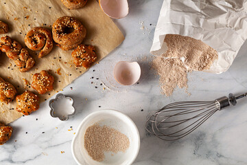 Flat lay image of  fresh handmade pastry cooling on baking paper with seeds scattered on marble kitchen countertop. A whisk, egg shells, wholewheat flour, measuring cup and cookie cutter are seen