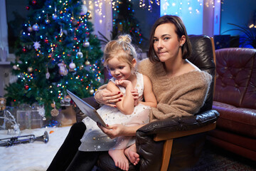 Atmospheric and shiny room with christmas tree and lights and little girl celebrating New Year with her mother sitting on leather armchair with tablet.