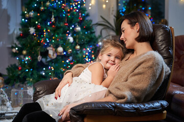 Joyful and happy little girl and her mother sitting on leather armchair together in cosy and atmospheric living room with christmas tree and lights.