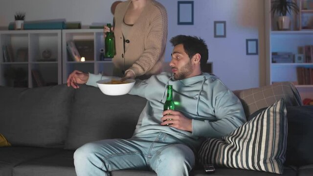 Tracking right of young Mixed-Race man dressed casually sitting on couch at home, holding bottle of beer, watching TV, red-haired Caucasian girlfriend bringing him bowl of snacks