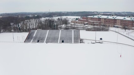 Aerial of school athletic fields and bleachers covered in winter snow. University college campus in United States of America during snowstorm. Aerial drone overhead view. - Powered by Adobe