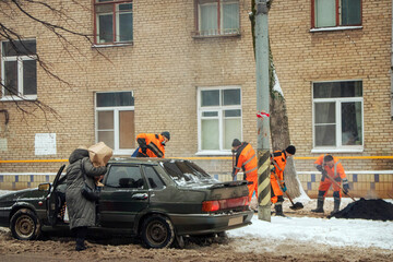 Workers in winter in snowy weather change the asphalt. The woman gets out of the car.