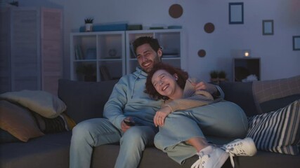 Tracking right of loving diverse couple cuddling on couch at home at night, smiling, cuddling, laughing at comedy on TV. Red-haired Caucasian girl leaning on chest of loving Mixed-Race boyfriend - Powered by Adobe