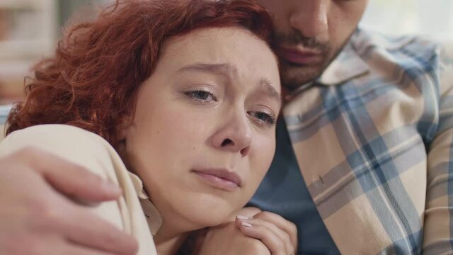Close-up of young blue-eyed Caucasian woman with copper hair weeping on chest of cropped partner in checkered shirt patting her on shoulder