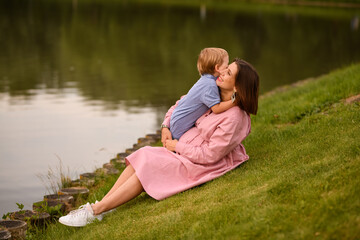 Woman with her son on the bank of the pond. They sit huddled. The calm water of the pond reflected scenic shore. Boy and woman admiring the scenery. Peace and harmony