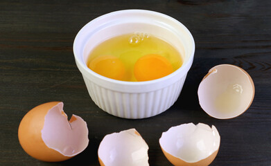Closeup two of raw eggs in a bowl on black wooden table with cracked eggshells in foreground