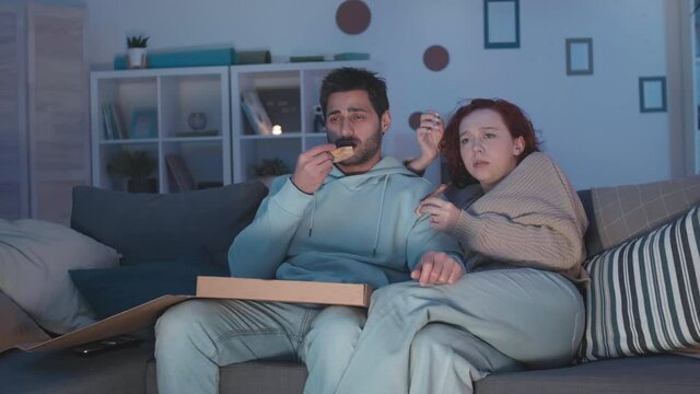 Tracking right of young Mixed-Race guy and red-haired Caucasian girl sitting in apartment on couch, enjoying pizza from box, watching, cringing and laughing at movie on TV at night