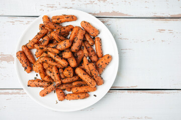 White plate with small peeled pieces of cooked baby carrot on white textured wooden table. Top view. Copy space.