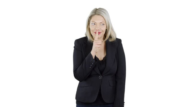 An attractive blonde haired woman is holding her finger to her lips gesturing to be quiet like keeping a secret. The girl is wearing a business suit with white screen background.