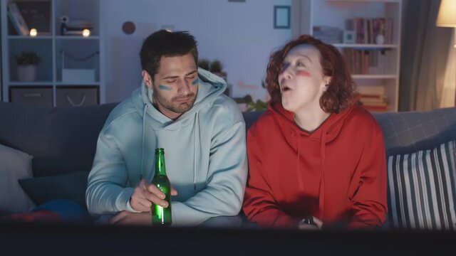 Medium shot of Caucasian woman in red celebrating favorite team, mocking, showing tongue to Mixed-Race man in blue with stripes painted on face, watching game on TV at night, holding bottles of beer