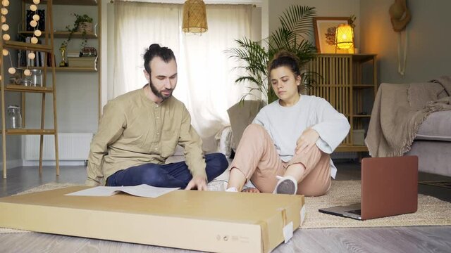 Millennial young couple looks at manual to assemble new furniture sitting on floor with cardboard box and modern laptop in light living room