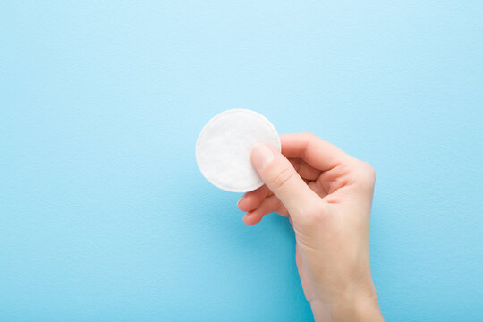 Young adult woman hand fingers holding white soft cleansing cotton pad on light blue table background. Pastel color. Closeup. Top down view.