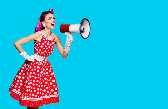 Violet red haired fashionable trendy woman using megaphone, shouting something. Girl in pin-up style dress in polka dot isolated over aqua blue color background. Retro fashion vintage studio concept.