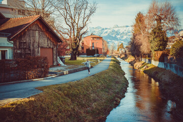 Happy girl in Alps mountains in Liechtenstein. Medieval Red House, calm narrow mountain river and jogging track, residential buildings, blue sky and snow-capped mountains. Liechtenstein, Vaduz