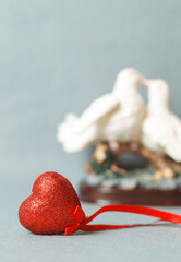 Red decorative shiny heart on a gray background. A statuette with kissing doves in the background. Copy space, selective focus