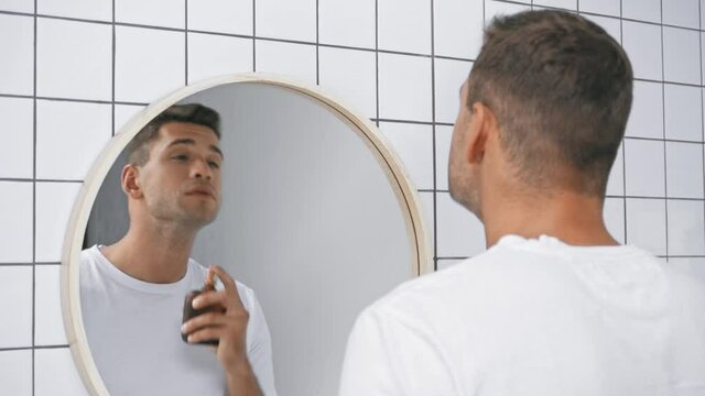 man in white t-shirt spraying perfume and looking at mirror