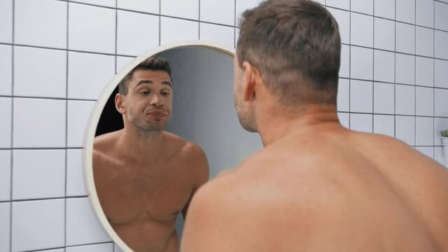 sleepy man yawning, touching face and puffing cheeks while looking at mirror