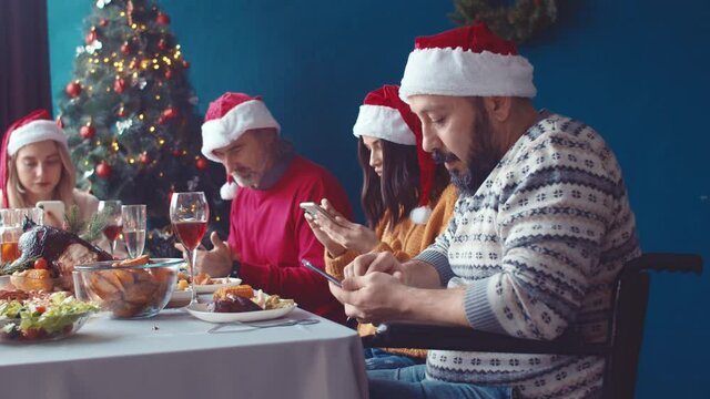 Family with man in wheelchair uses phones during Christmas holiday. Group of friends use their smartphones at festive dinner table. Dependence on phone. Nomophobia