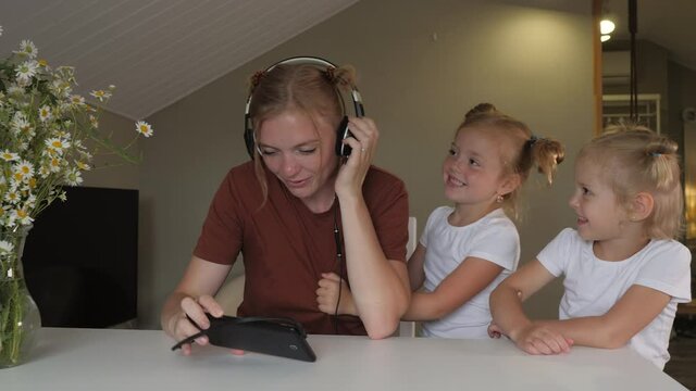 Young mother tries to work remotely at home while two naughty little daughters distract her. Two fidgety sisters pester their mom while she works with her tablet and headphones.