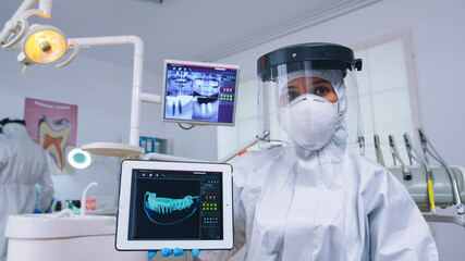 Fototapeta na wymiar Doctor showing digital x-ray on tablet in dental office with new normal, explaining teeth treatment. Stomatology wearing protective suit against infection with coronavirus pointing at radiography.