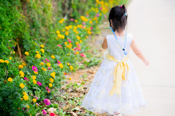 Obraz na płótnie Canvas Selective focus. Back view of girl in a beautiful dress is walking on the street. Along the street road, planted colorful flowers beautiful. Children walk to see flowers. Kid wearing medical face mask