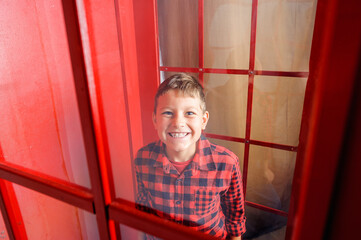 a cheerful smiling fair-haired teenage boy, dressed in a red and black plaid shirt, stands in a red telephone box