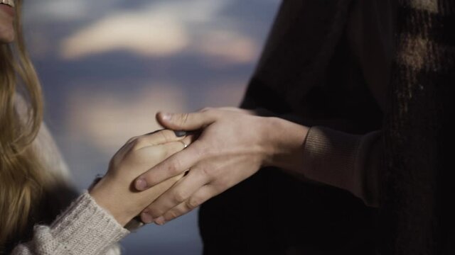 Close-up of unrecognizable boyfriend caressing hands of smiling young girlfriend with beautiful evening sky at the background. Romantic Caucasian loving couple dating outdoors at dusk. Unity concept.