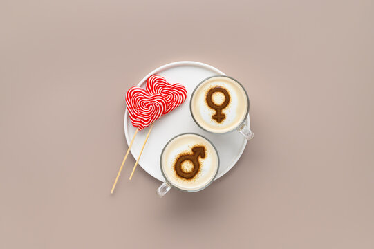 Two cups of coffee with symbols of mars and venus on whipped milk foam and lollipops in heart shape on white plate. Beige background. Concept romantic date on Valentine's day. Flat lay