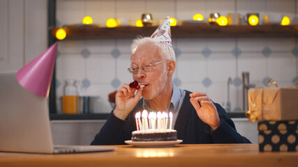 Senior man in cap blowing party whistle holding birthday cake and waving hand having video call