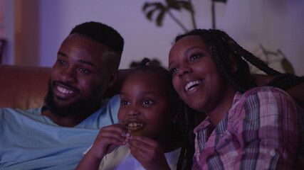 Close up of happy african mother, father and daughter watching tv on couch at home
