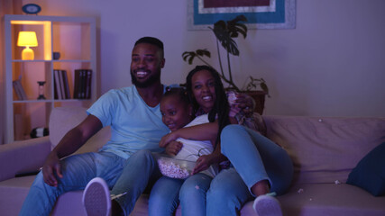 Afro-american mom, dad and daughter watching sports game on tv and eating popcorn