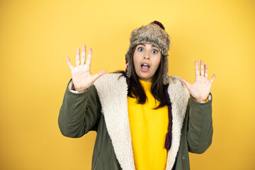 Young beautiful woman wearing a hat and a green winter coat over yellow background afraid and terrified with fear expression stop gesture with hands, shouting in shock. Panic concept.