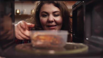 Beautiful young woman opening microwave oven and taking plastic container with heated meal