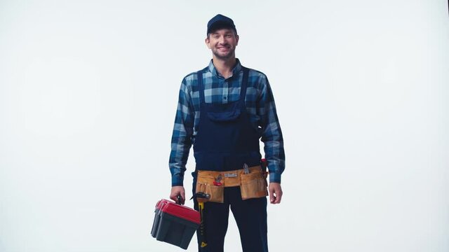 happy plumber in uniform adjusting cap and holding toolbox on white