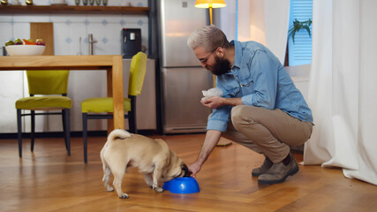 Young man feeding pet dog at home offering it bowl of food