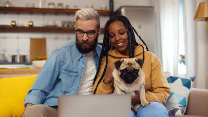 Happy multiethnic couple relax with dog on couch and having video call on laptop