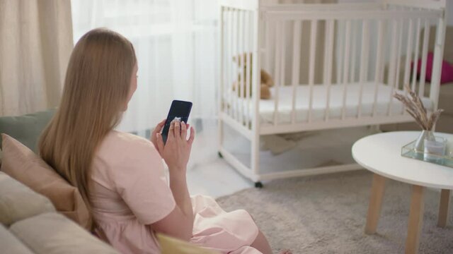 Medium back-view shot of unrecognizable woman sitting alone in bright living room looking at photo of baby ultrasound with white rocking cradle in background