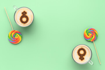 Two cups of coffee with symbols of venus on whipped milk foam and couple round LGBT rainbow lollipops on Pastel green ash color tabletop. Concept lesbian love. Flat lay, top view, copy space