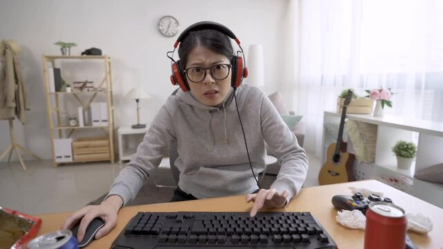 concentrated young gamer woman in headphones and glasses using computer for playing game at home. asian chinese female face camera as looking at monitor. nervous girl geek having fun online frowning