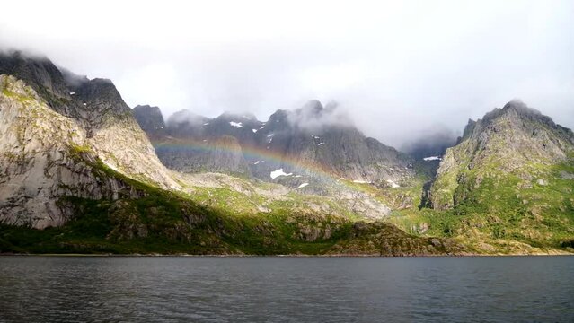Boat cruise on Raftsundeet strait in front of Austvågøya island, Lofoten, Norway with majestic rugged mountains covered by green plants and beautiful colorful rainbow on cloudy day in late summer.