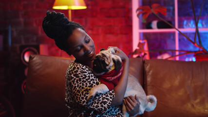 Fototapeta na wymiar Affectionate young afro woman hugs dog with love and care sitting on couch at home